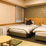 Hotel Dormy Inn Kumamoto – Affordable Stay with Onsen Comfort A minimalist room at Hotel Dormy Inn Kumamoto features two single beds with star-shaped pillows, a round wooden table with green floor cushions, paper sliding window, beige walls, and a small air purifier.