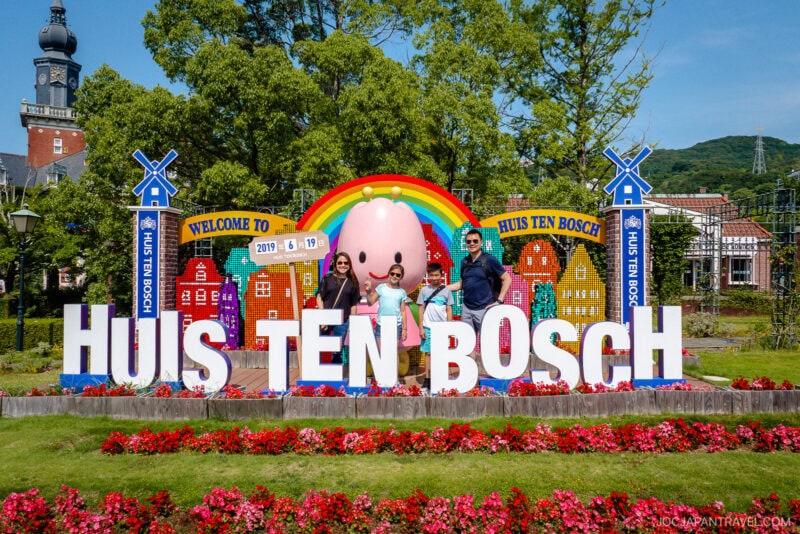 Three people pose in front of a colorful Huis Ten Bosch sign with a large mascot, rainbow, windmills, and Dutch-style buildings in the background, surrounded by flowers and greenery under the bright blue sky at Huis Ten Bosch.