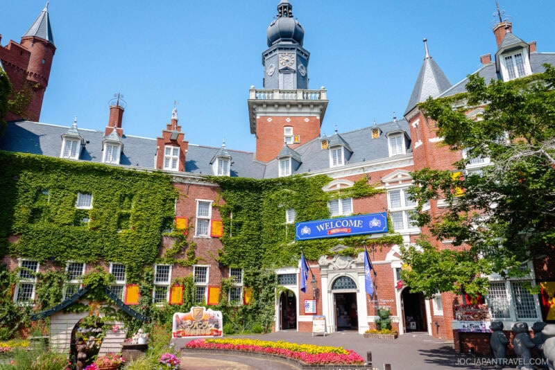 A large, ivy-covered brick building with a tall clock tower and pointed turrets evokes the charm of Huis Ten Bosch. A blue Welcome banner hangs above the main entrance, while bright flowers and greenery decorate the lively courtyard.