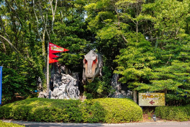 A life-sized dinosaur model with an open mouth stands among greenery and rocks, next to a red flag and a sign with Japanese text, welcoming visitors at the Huis Ten Bosch theme park entrance.