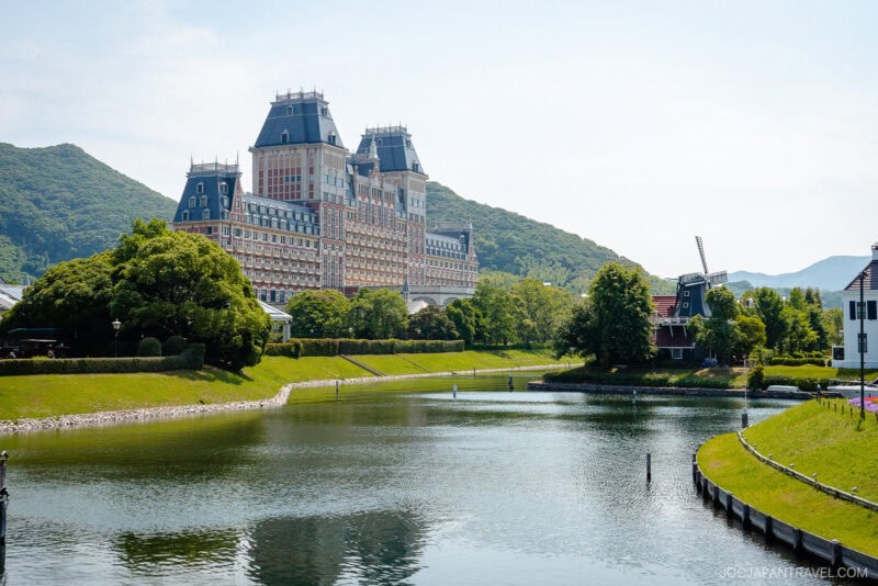 A large European-style building with a mansard roof stands beside a calm river in Huis Ten Bosch, surrounded by green trees, with hills in the background and a windmill on the right.
