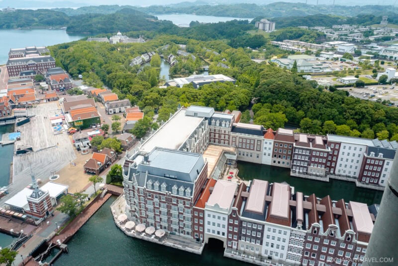 Aerial view of Huis Ten Bosch, a European-style resort with canals, red-roofed buildings, and lush greenery, surrounded by water and a distant hilly landscape under a cloudy sky.