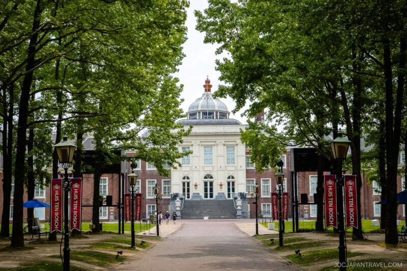 A grand white building with a domed roof stands at the end of a tree-lined path in Huis Ten Bosch, flanked by brick buildings and red banners. The scene is serene, with green foliage and a few people walking nearby.
