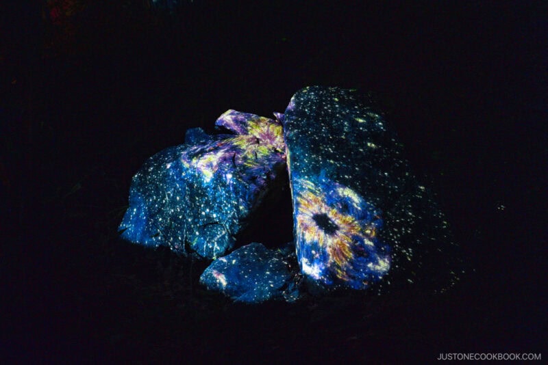 Large rocks at Jozankei Onsen are illuminated with colorful, galaxy-like projections featuring star and nebula patterns, set against a dark background.