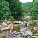 Jozankei Onsen (定山渓温泉) – A Hot Spring Retreat Near Sapporo Two children stand on rocks in a shallow, clear river surrounded by lush green trees near Jozankei Onsen. A red bridge spans the background with forested hills behind it. The peaceful scene is bright and serene.