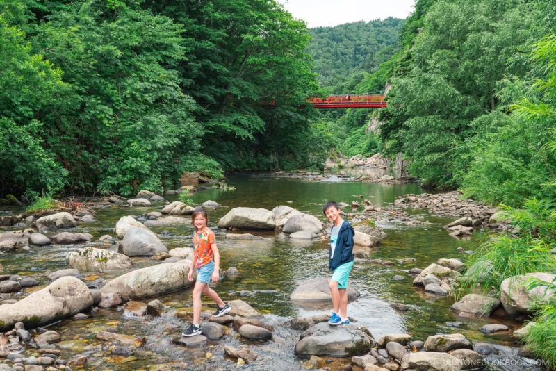 Two children stand on rocks in a shallow, clear river surrounded by lush green trees near Jozankei Onsen. A red bridge spans the background with forested hills behind it. The peaceful scene is bright and serene.