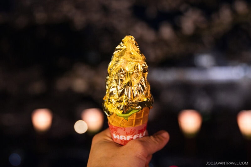 A hand holds an ice cream cone covered in shiny gold foil against a blurred night background with warm lights and cherry blossoms, offering a glimpse into the luxurious treats featured in the Kanazawa food guide.