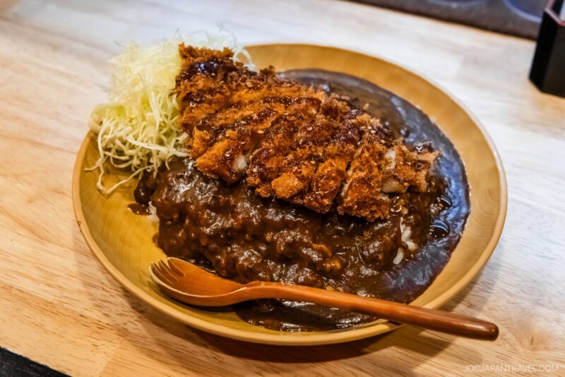 A wooden plate with Japanese katsu curry—breaded, fried pork cutlet atop rice covered in dark curry sauce, served with shredded cabbage and a wooden spoon—perfect for any Kanazawa food guide.