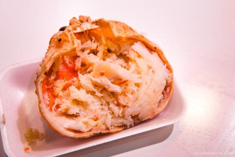 A close-up of cooked crab meat served in a halved crab shell on a white tray, showcasing juicy, shredded crab flesh and a bit of orange roe—a must-try delicacy featured in any Kanazawa food guide.