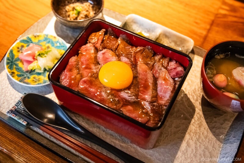 A Japanese meal featured in the Kanazawa food guide: grilled beef topped with raw egg yolk, sashimi, pickles, soup, and side dishes—all artfully arranged on a tray with chopsticks and a spoon.