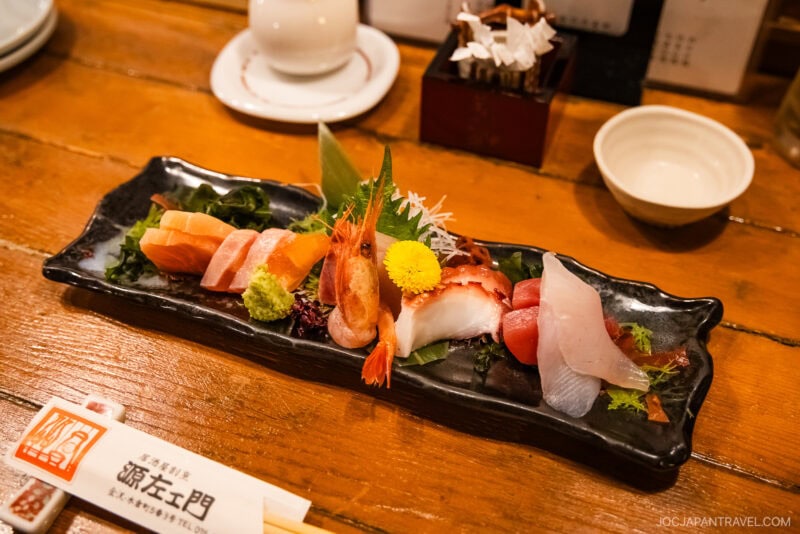 A black rectangular plate with assorted sashimi, including shrimp, tuna, salmon, and white fish—an elegant presentation featured in the Kanazawa food guide—garnished with wasabi, greens, and a yellow flower on a wooden table with chopsticks and dishes.