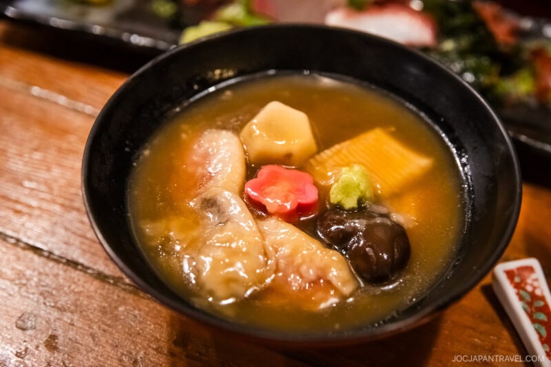 A black bowl filled with Japanese soup—featuring various vegetables, mushrooms, and fish, including a red flower-shaped slice—served on a wooden table. A must-try from the Kanazawa food guide.