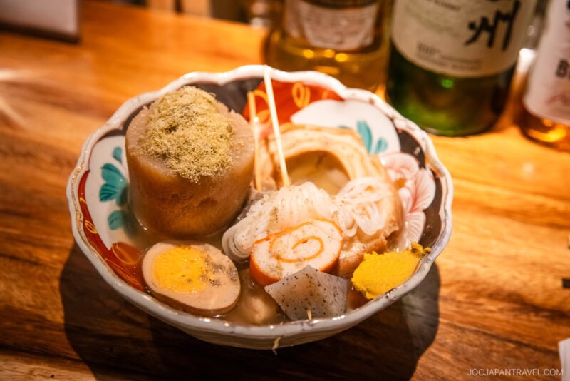 A colorful bowl of Japanese oden with boiled egg, daikon radish, fish cakes, shirataki noodles, and mustard on a wooden counter&mdash;an essential highlight in any Kanazawa food guide&mdash;with bottles in the background.