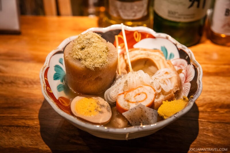 A colorful bowl of Japanese oden—daikon radish, boiled egg, fish cakes, konjac, shirataki noodles, and mustard in light broth—sits on a wooden table, a must-try in any Kanazawa food guide for authentic regional flavors.