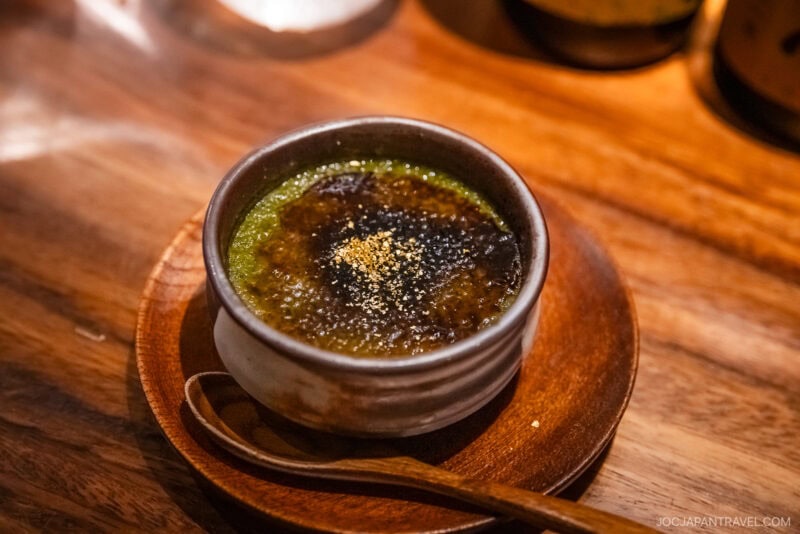 A ceramic bowl of matcha crème brûlée with a caramelized sugar crust, served on a wooden plate with a wooden spoon, awaits on a polished table—an elegant highlight in any Kanazawa food guide.