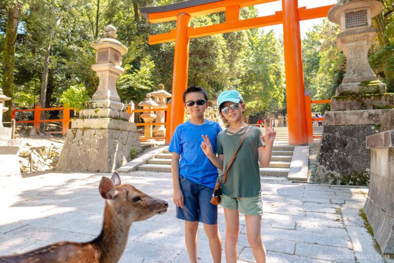 Two children pose for a photo near a deer in front of a bright orange torii gate at Kasuga Taisha, surrounded by stone lanterns and lush green trees on a sunny day.