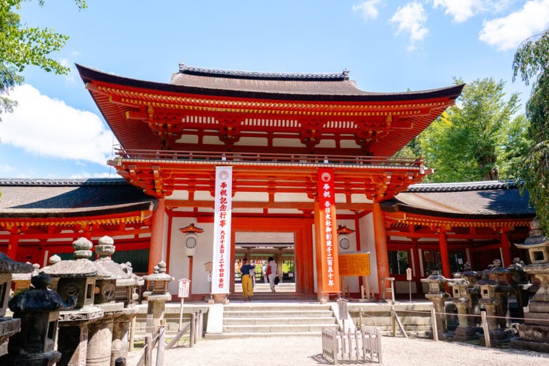 A traditional Japanese shrine entrance, reminiscent of Kasuga Taisha, features a large red gate, white banners with Japanese writing, stone lanterns on each side, and a bright blue sky in the background.