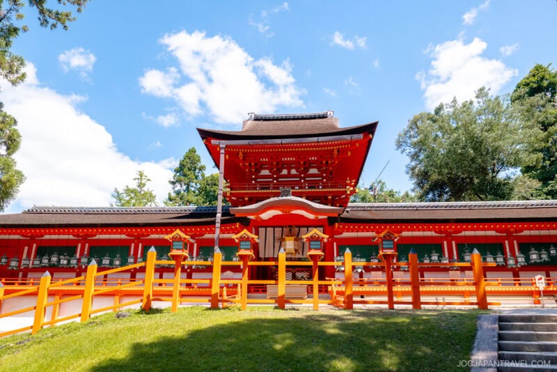 A bright red Shinto shrine inspired by Kasuga Taisha, with ornate details and lanterns, stands under a sunny blue sky, surrounded by green trees and a grassy foreground.