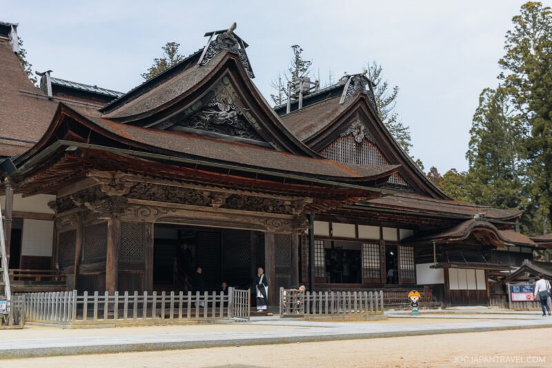 A traditional Japanese wooden temple in Koyasan with ornate, curved roofs, intricate carvings, and a fenced veranda surrounded by trees. Two people are visible near the entrance.