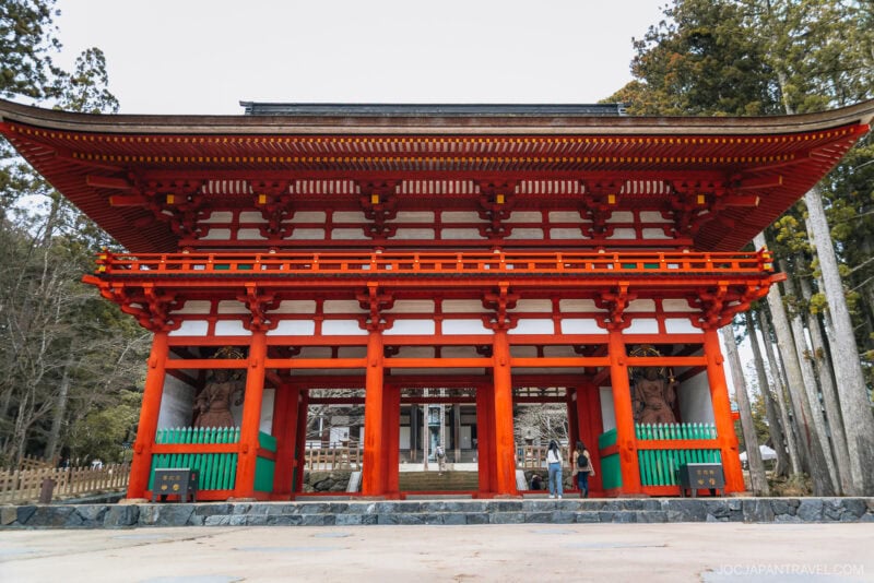 Large traditional Japanese torii gate painted red with white and wooden accents stands at the entrance to a koyasan shrine or temple, with people walking underneath and trees in the background.