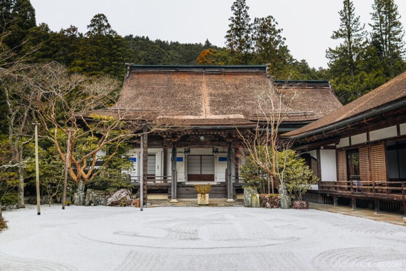 A traditional Japanese temple with a thatched roof stands behind a peaceful Zen garden featuring white gravel patterns, surrounded by trees and wooden walkways in the serene mountain town of Koyasan.