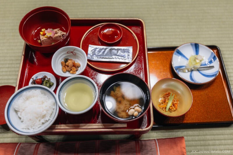 A traditional Japanese meal inspired by Koyasan, served on a lacquer tray with rice, miso soup, pickled vegetables, green tea, simmered beans, sashimi, a soy sauce packet, and garnishes—all beautifully arranged in small bowls and plates.