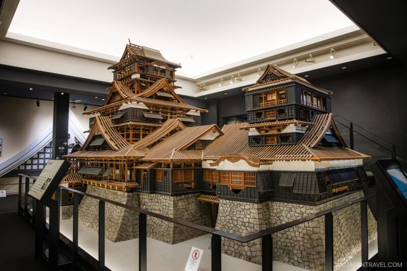 A detailed scale model of Kumamoto Castle is displayed indoors, surrounded by glass barriers. The model highlights wooden structures, tiled roofs, and stone walls, all illuminated by soft overhead lighting.