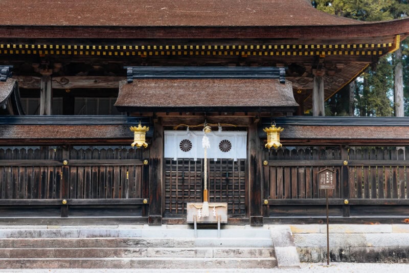 A traditional Japanese wooden shrine inspired by Kumano Hongu Taisha, with a thatched roof, white doors featuring two black symbols, golden lanterns, and a rope in front for ceremonial purposes.