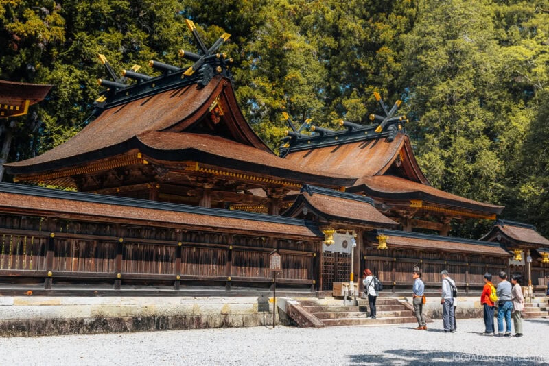 A traditional Japanese shrine, Kumano Hongu Taisha, with ornate wooden roofs and gold accents stands among tall trees, while five people stand outside taking photos and admiring the architecture on a sunny day.