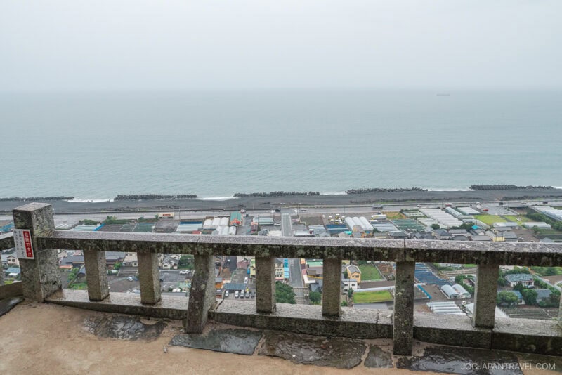View from a stone balcony overlooking a coastal town with colorful rooftops, reminiscent of the vistas near Kunozan Toshogu Shrine, a road winding below, and the ocean stretching to the horizon under an overcast sky.