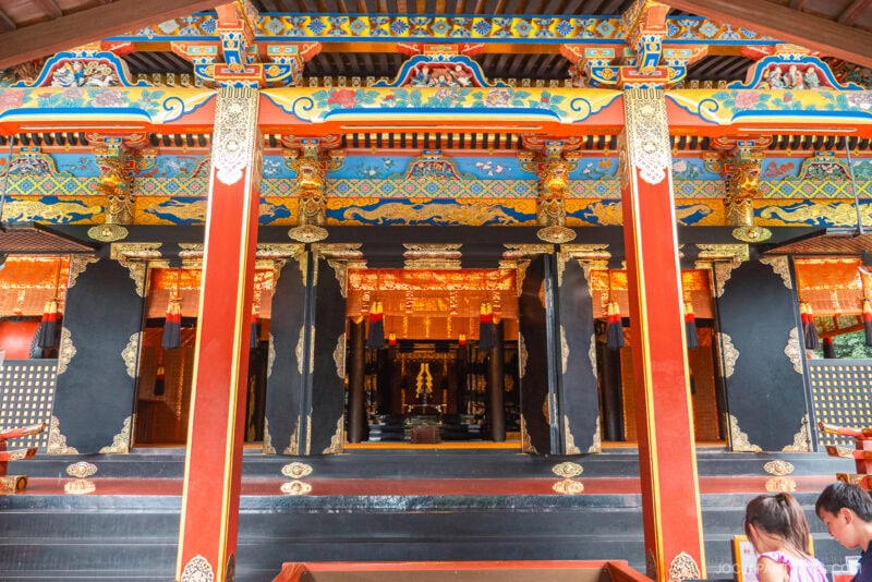 Vibrantly painted and ornately decorated Kunozan Toshogu Shrine entrance with gold details, red pillars, and intricate patterns; two people are partially visible in the bottom right corner.