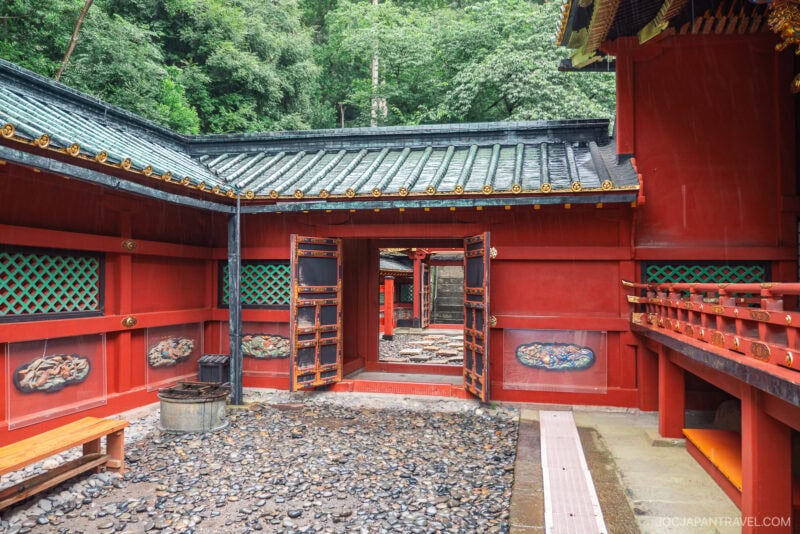 A traditional Japanese shrine courtyard, inspired by Kunozan Toshogu Shrine, with red wooden walls, green lattice windows, and an open gate leading outside. The ground is covered with stones and surrounded by lush green trees.