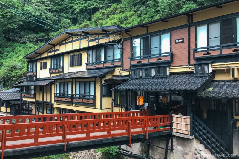 A traditional Japanese inn with yellow and brown wooden walls stands beside a lush green hillside in Kurokawa Onsen. A bright red bridge crosses over a small stream in front of the building.