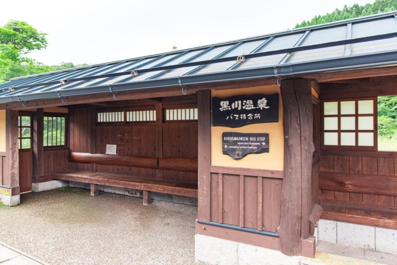 A wooden bus stop shelter with benches and a sign that reads Kurokawa Onsen Bus Stop, nestled amid lush greenery and mountains near the famed Kurokawa Onsen hot spring area.
