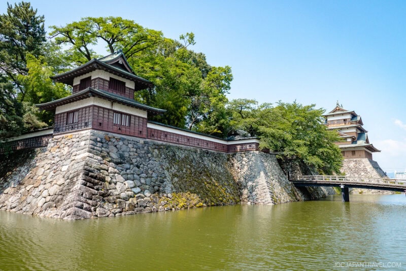 Traditional Japanese castle buildings with stone walls stand beside a calm moat, surrounded by lush green trees under a clear blue sky. A wooden bridge crosses the water to the entrance, reminiscent of historic castles near Lake Suwa.
