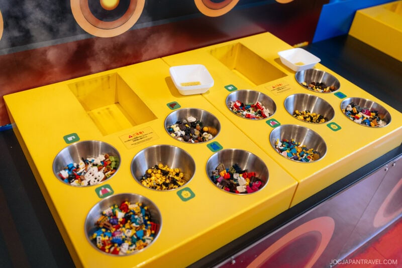A yellow sorting table with metal bowls filled with various LEGO pieces in different colors, and small white trays for collecting selected pieces—an interactive setup you might find featured in a Legoland Nagoya travel guide.