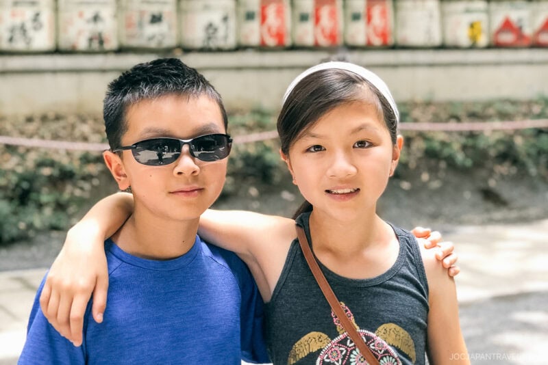 Two children stand close together outdoors near Meiji Jingu Shrine, one wearing sunglasses and a blue shirt, the other with a headband and dark tank top, smiling with an arm around their friend’s shoulders. A blurred wall and greenery are in the background.