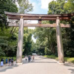 Meiji Jingu Shrine (明治神宮) – A Peaceful Forest Retreat in Tokyo Visitors walk under a large wooden torii gate surrounded by tall green trees on a wide path at Meiji Jingu Shrine in Tokyo, Japan, on a sunny day.