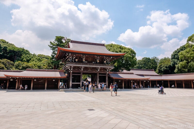 Large traditional Japanese shrine with a wooden gate and sloped roof, surrounded by trees. Visitors stroll across the spacious stone courtyard of Meiji Jingu Shrine beneath a partly cloudy sky.