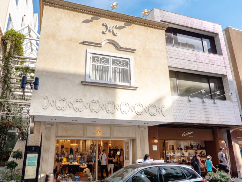 A light-colored, two-story shop with ornate exterior designs and a large window above the entrance sits along Motomachi Shopping Street. People are entering and exiting the store, while a black car is parked in front on a sunny day.