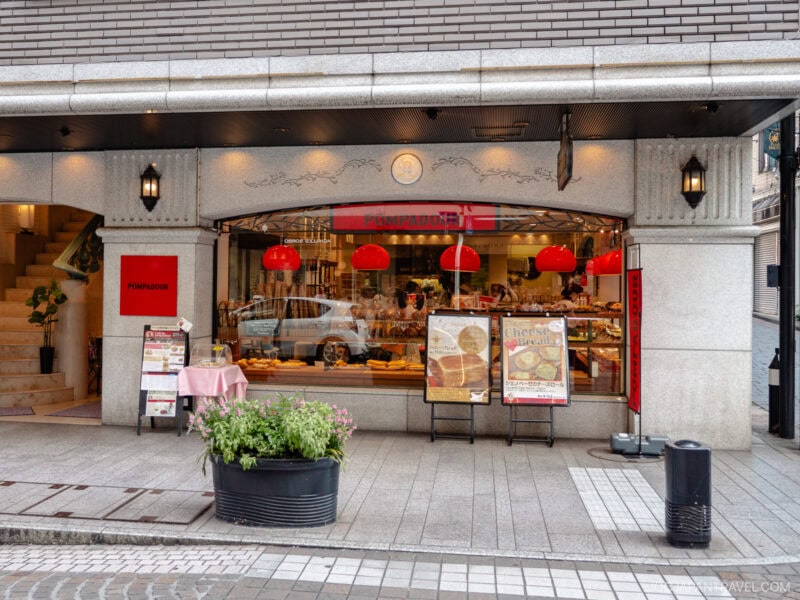 A bakery storefront on Motomachi Shopping Street features large windows displaying breads and pastries. Red pendant lights hang inside, while two menu boards, a table with a pink cloth, and a flower planter sit outside by the entrance.