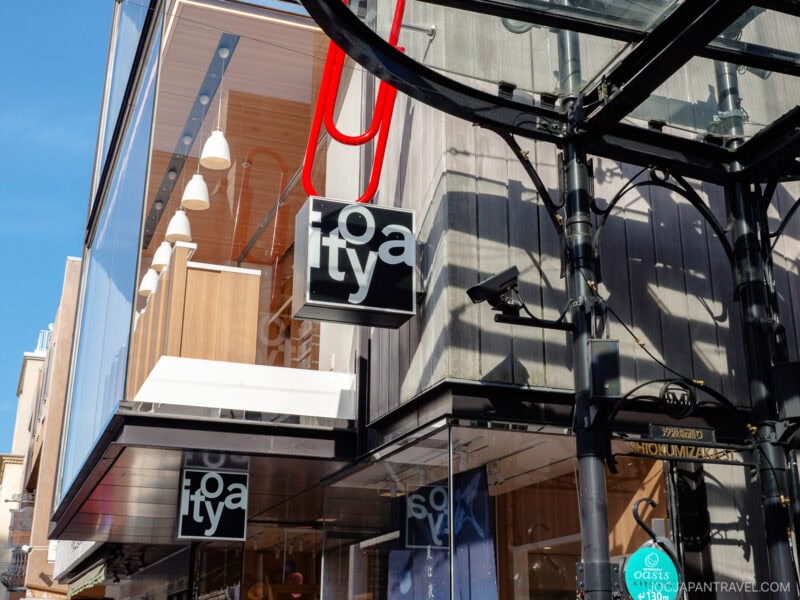 A modern storefront with glass and wood elements features two black and white lofty signs. Bright sunlight casts shadows, and a blue sky is visible above. The reflection of Motomachi Shopping Street can be seen in the glass.