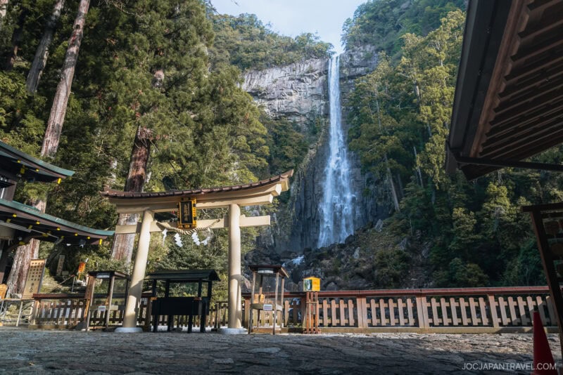 A tall waterfall, Nachi Falls, cascades down a forested cliff behind a traditional Japanese torii gate and shrine structures, surrounded by tall trees and stone paving.