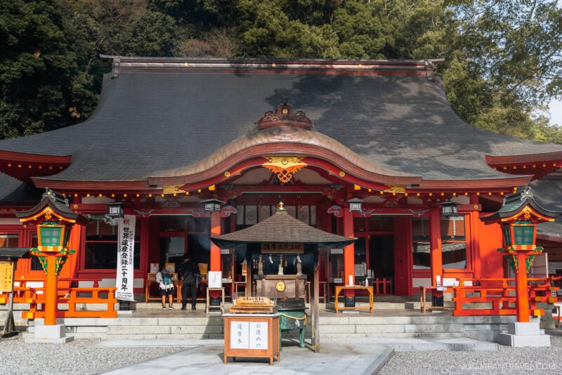 A traditional Japanese shrine with a curved roof, bright red-orange pillars, lanterns, and wooden plaques at the entrance, nestled near lush greenery and Nachi Falls, with a few visitors gathered by the steps.