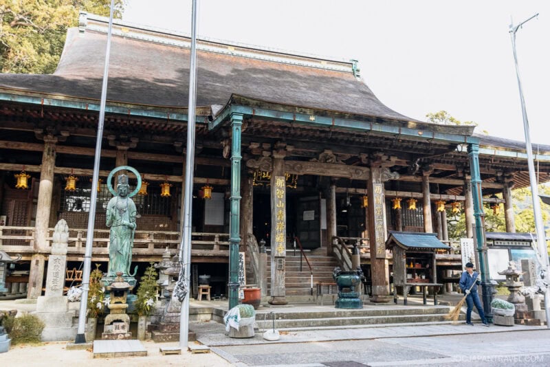 A traditional Japanese temple with wooden architecture and a sloping roof, reminiscent of those near Nachi Falls. A person sweeps the ground by a large green standing statue as lanterns hang from the eaves and various small structures surround the scene.