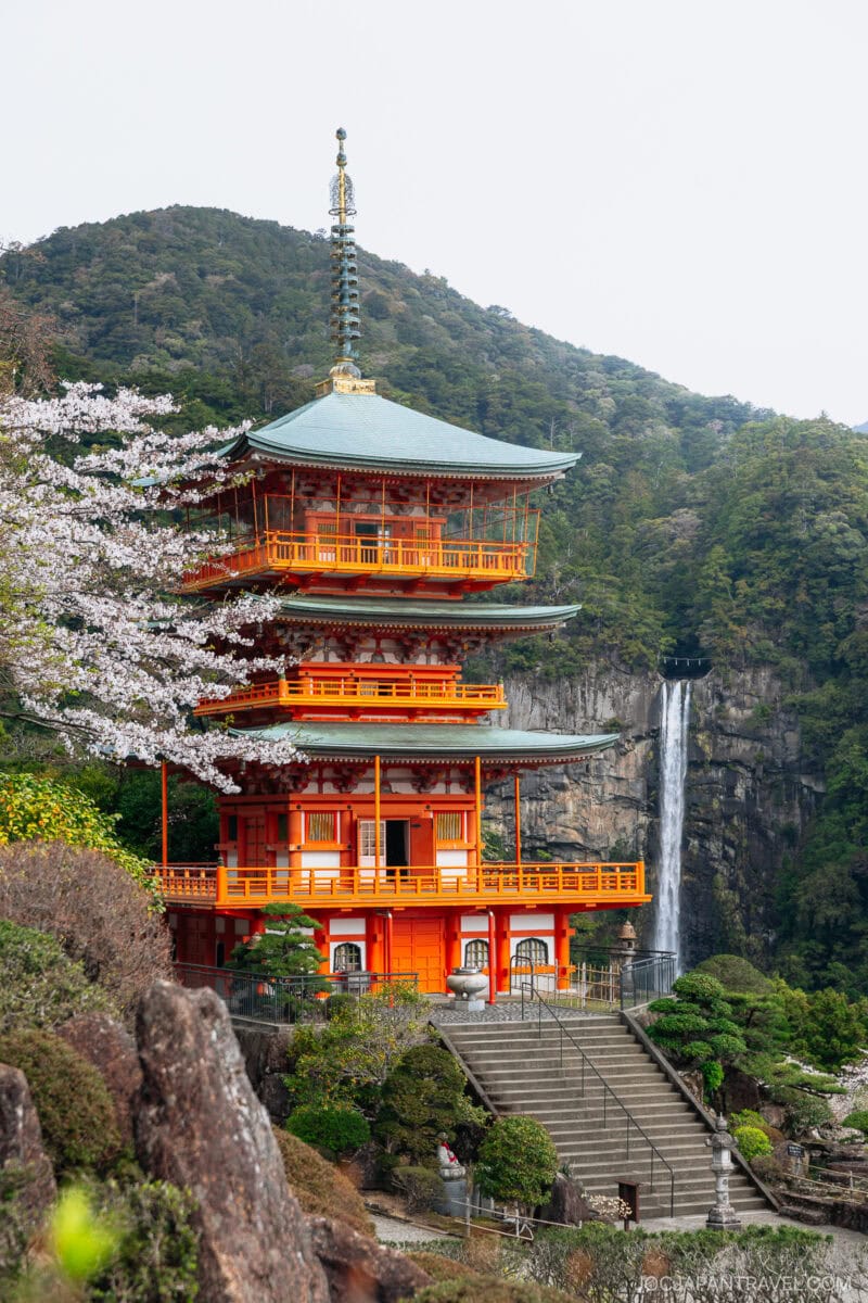A vibrant red three-story pagoda stands amid lush greenery and cherry blossoms, with the majestic Nachi Falls cascading down a cliff in the background.