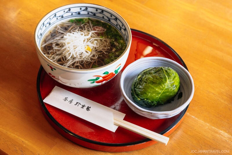 A bowl of soba noodle soup topped with sliced green onion, served with a green, leaf-wrapped rice ball reminiscent of treats enjoyed near Nachi Falls, and chopsticks on a red tray on a wooden table.