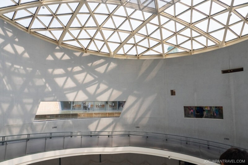 A large, curved interior wall under a glass dome casts geometric shadows at the Nagasaki Atomic Bomb Museum, with two rectangular windows displaying art and a few people visible inside the modern museum space.