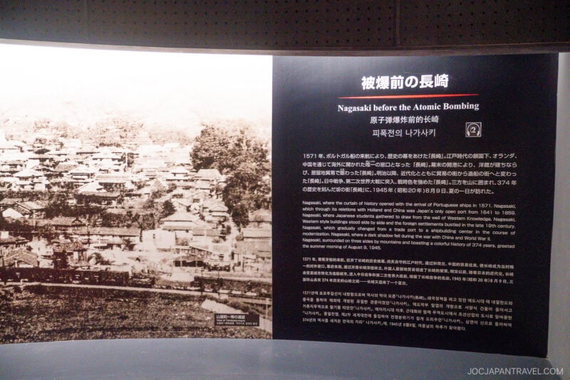 A display panel at the Nagasaki Atomic Bomb Museum shows a historic photo of Nagasaki before the bombing, with Japanese, English, Korean, and Chinese text detailing the city’s history and pre-war context.