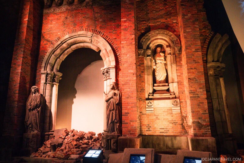 A brick wall at the Nagasaki Atomic Bomb Museum features a large arched doorway, two standing stone statues, a pile of rubble, and a statue of a robed figure in a niche. The dim lighting creates a dramatic atmosphere.